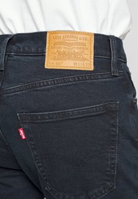 Jeans de denim oscuro con un parche de cuero de la marca en la parte posterior de la cinturilla, costuras de bolsillos traseros en curva y una pequeña etiqueta roja en el bolsillo.