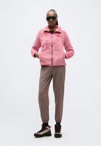 Γυναίκα στέκεται με τα χέρια στις τσέπες, φορώντας ροζ υφασμάτινο μπουφάν με φερμουάρ, παντελόνι τζόγκινγκ σε χρώμα ταούπε, ορειβατικά παπούτσια και μεγάλα ανακλαστικά γυαλιά ηλίου.