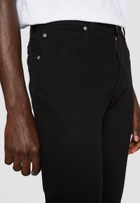 Personne à la peau foncée portant un jean noir ajusté et une chemise blanche, montrant la poche latérale et la main reposant près de la cuisse.