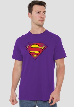 Young man wearing a purple T-shirt with a distressed red and yellow Superman logo, looking to his left against a plain light background.