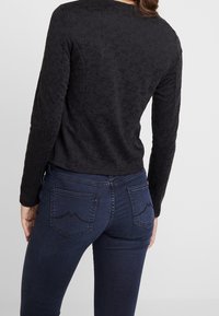 Černý top s dlouhým rukávem s texturovaným vzorem, kombinovaný s tmavě modrými džínami s zakřivenými zadními kapsami. Materiál je hladký a přiléhavý.