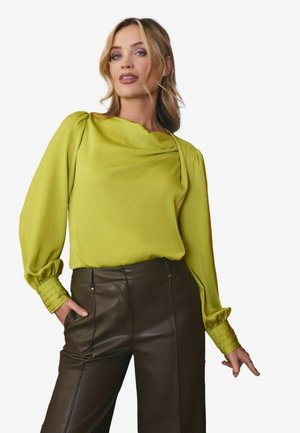 Blusa verde lime con scollatura drappeggiata e maniche ampie, abbinata a pantaloni in pelle marrone con cuciture laterali e dettagli delle tasche.