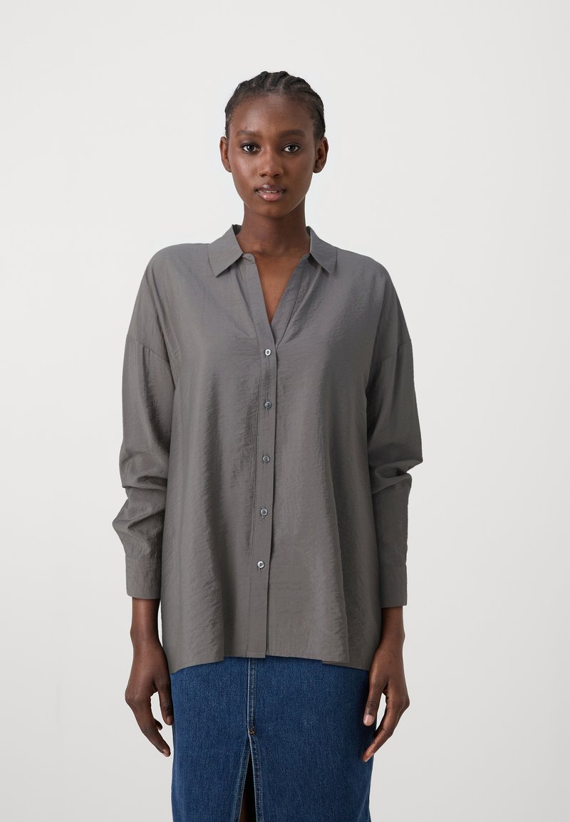 Camisa cinza de botões, feita de tecido leve, com gola, mangas longas e um corte relaxado. Combinada com uma saia de ganga.