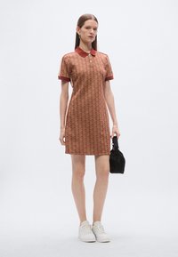 Minta mintás barna anyagból készült polo ruha piros gallérral és ujjakkal. Vékony szabású és enyhén texturált felületű. Fehér tornacipő és fekete táska.