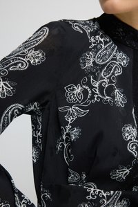 Schwarze Bluse mit weißen Blumen- und Paisley-Mustern, transparentem Stoff und Spitzenverzierung am hohen Kragen, getragen von einer Person mit angewinkeltem Arm.