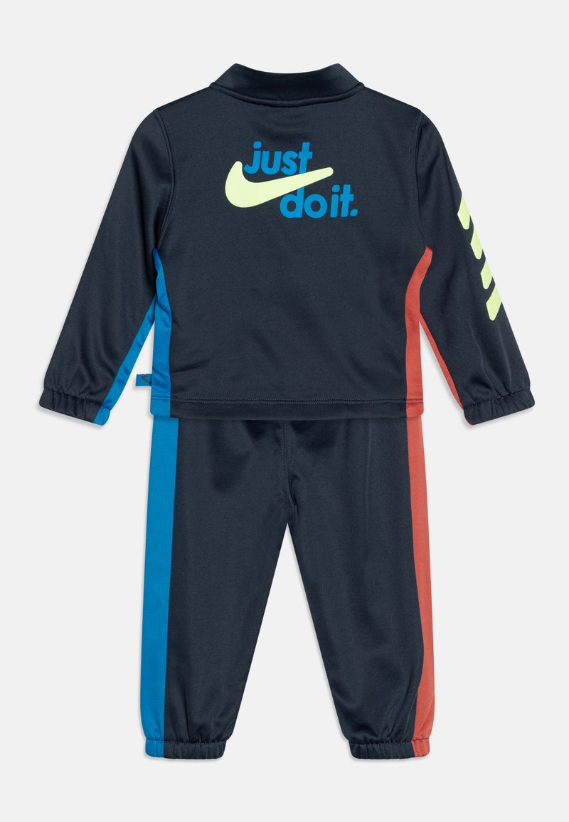 Chándal azul marino que presenta una chaqueta con cuello y el logo verde lima "Just Do It", así como franjas laterales en azul brillante y rojo en los pantalones.