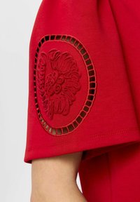 Haut rouge à manches courtes avec un design floral brodé sur la manche, présentant des découpes et une texture de tissu lisse.