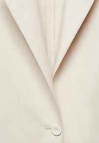 Béžové sako s klopovým límcem, dvěma předními záševky a zapínáním na jeden bílý knoflík. Hladká látka s lesklou texturou.