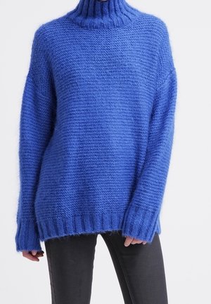 Løs, blå strikket rullekrave sweater båret med sorte bukser, der viser tekstur og ribbekanter på ærmer og kant.