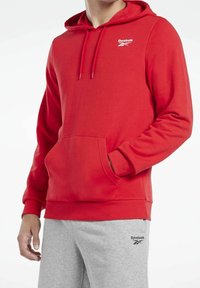 Haut à capuche rouge Reebok avec une poche frontale, un cordon de serrage, des poignets côtelés et un logo sur la poitrine. Tissu doux avec une texture lisse.