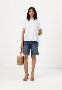 Hvit peplum-top kombinert med blå denimshorts. Modellen holder en vevd stråveske. Lysebrune stroppesandaler fullfører antrekket.