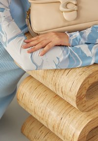 Beige läderhandväska med en unik lås som vilar på en böjd, skiktad träram. Blå mönstrad tygärm synlig med delikata texturer.