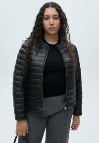 Jachetă de tip puffer neagră cu textură matlasată, închidere cu fermoar și manșete ribbate. Pur dată peste un top negru, asortată cu pantaloni gri cu dungi.