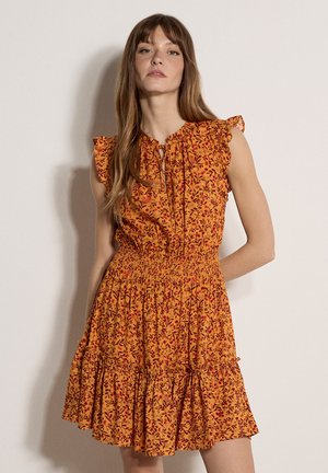 Femme portant une robe orange sans manches à motifs floraux, avec des manches à volants et une taille froncée, debout devant un fond clair uni.