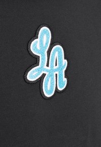 Tela negra con un destacado logo bordado "LA" en azul claro y blanco, mostrando una fuente estilizada y juguetona.