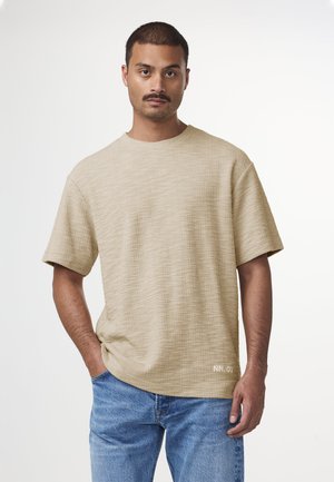 Beige T-shirt met korte mouwen van textuurstof, ronde halslijn en subtiele geborduurde details aan de zoom. Draagt met blauwe jeans.