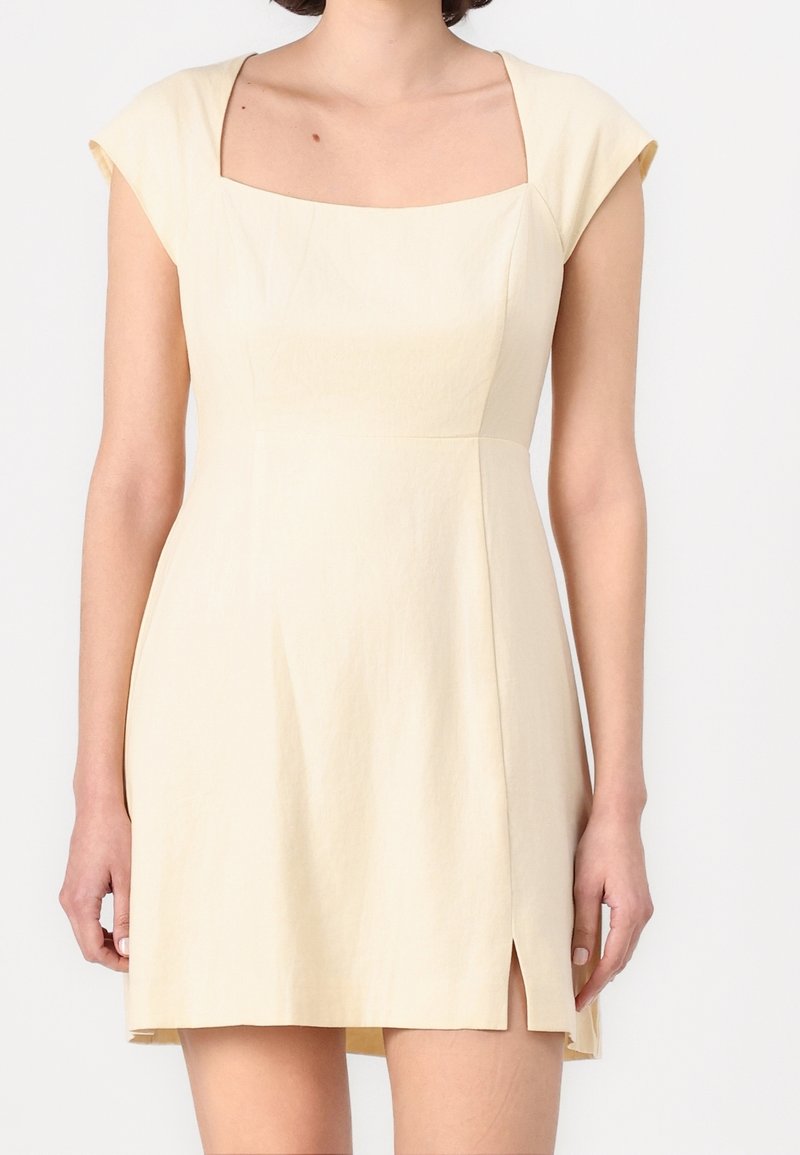 Femme portant une robe courte beige clair avec des manches courtes, un col carré et une petite fente sur le côté gauche.