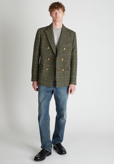 Giacca doppiopetto verde a quadri con bottoni dorati, abbinata a una t-shirt grigia e jeans blu. Le scarpe nere completano l'outfit.