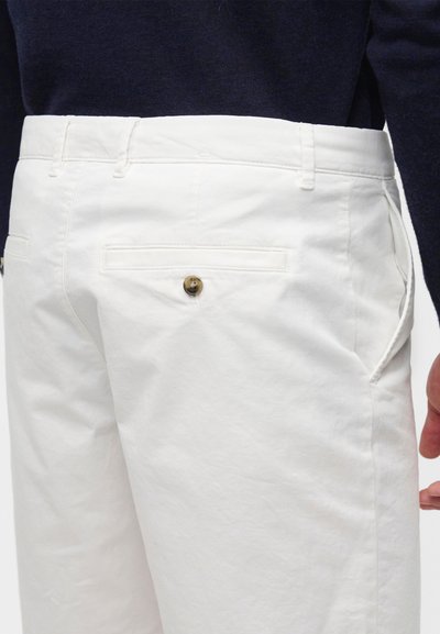 Gros plan d'une personne portant un pantalon blanc avec une poche arrière boutonnée, une poche latérale et un haut foncé visible à la taille.