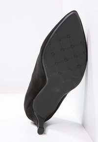 Scarpa nera in suede con punta affilata e tacco kitten, inclinata per mostrare la suola in gomma a forma di cuore, su sfondo bianco.