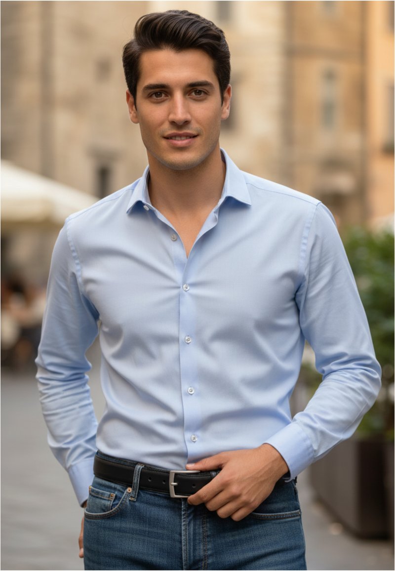 Joven con camisa azul clara y vaqueros de pie al aire libre, ajustándose el cinturón negro, con fondo urbano desenfocado.