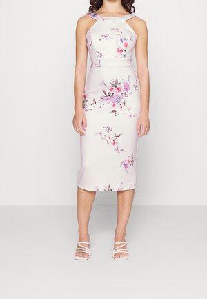 Kvinde iført en ærmeløs, hvid midi-kjole med pink og lilla blomsterprint og hvide sandaler med stropper står foran en ensfarvet baggrund.