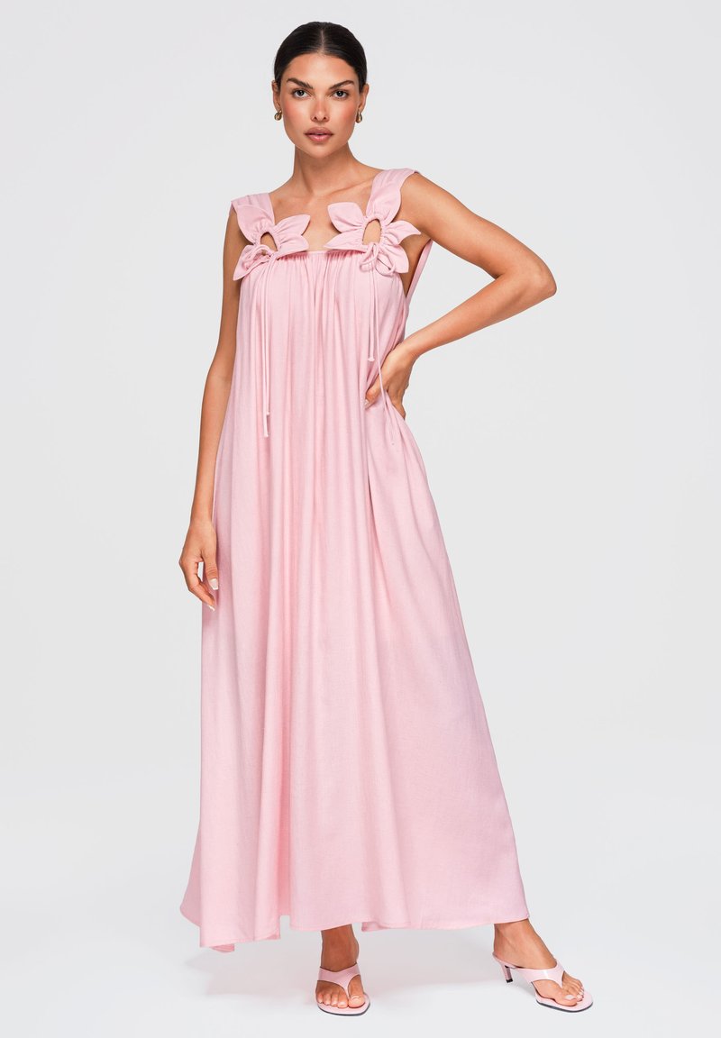 Femme portant une longue robe rose sans manches avec de grands détails floraux aux épaules et des sandales roses à talons, debout avec une main sur la hanche.