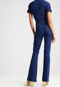 Denimbyxdress med korta ärmar, mörkblå färg, åtsittande midja, utsvängda ben och bakfickor; har logotyp vid halsringningen.