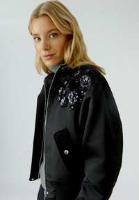 Schwarze Bomberjacke mit floralem Schmuck auf der linken Schulter, ausgestattet mit einem Reißverschluss, Taschen und gerippten Bündchen. Glatte Textur.