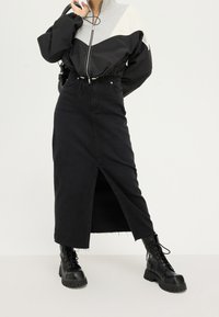 Zwarte denim maxi-rok met een vooraan gespleten plooi, gecombineerd met een zwart-grijs kleurgeblokt jack. De outfit wordt compleet gemaakt met robuuste zwarte combatlaarzen.