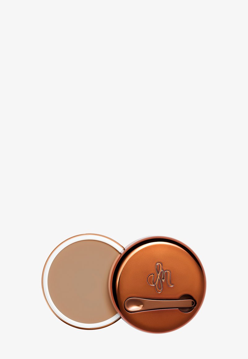 Ronde compact met een glanzende koperen deksel en gegraveerd logo, met een crèmekleurig product binnenin en een klein applicatortje.