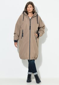 Beige wasserdichter Mantel mit Kapuze, Frontreißverschluss, seitlichen Taschen und elastischen Bündchen. Getragen über dunklen Jeans und schwarzen Stiefeletten.