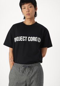 Črna bombažna majica s poudarjenim belim napisom "PROJECT CORE©." Okrogli ovratnik, kratki rokavi, v kombinaciji s sivo teksturiranimi hlačami.
