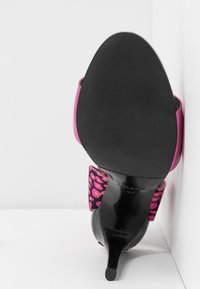 Chaussure à talon haut noire avec une bride en imprimé léopard rose et des côtés violets, photographiée par le dessous sur fond blanc.