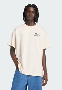 Béžové oversized tričko z bavlny, s kulatým výstřihem a krátkými rukávy, s černým logem Adidas na levé straně hrudi.