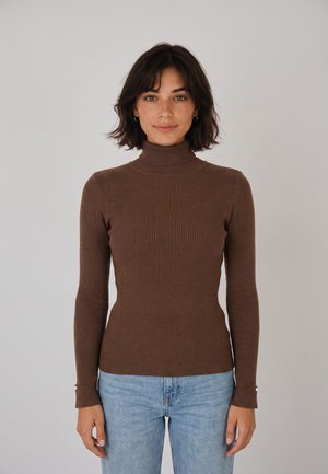 Donna con capelli corti e scuri che indossa un maglione marrone a collo alto a coste e jeans azzurri, in piedi davanti a uno sfondo chiaro uniforme.