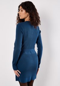 Mujer con cabello rizado oscuro que lleva un vestido azul de manga larga ajustado y con canalé, de pie de lado contra una pared blanca lisa.