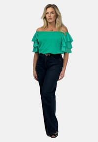 Zielony top bez ramiączek z warstwowymi falbanami, połączony z ciemnymi dżinsami o wysokim stanie. Stylizacja obejmuje pasek i sandały na obcasie odkrywające palce.