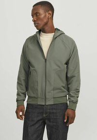 Chaqueta verde oliva con cremallera y capucha, con puños acanalados, un acabado suave, y dos bolsillos laterales, llevada sobre una camiseta de color claro.