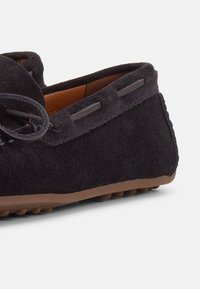 Μοκασίνι από μαύρο σουέντ με δερμάτινη λεπτομέρεια κορδονιού και καφέ λαστιχένια σόλα με αντιολισθητικές τελείες, εμφανιζόμενο από πλευρική γωνία.