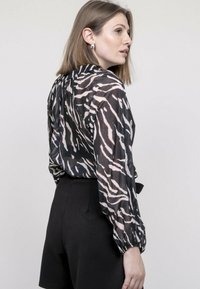 Blouse noire transparente avec un motif zèbre en blanc et rose pâle. Dotée de manches bouffantes et d'un détail noué dans le dos. Associée à un short noir taille haute.