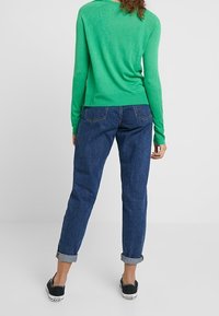 Maglione verde a maglia con maniche lunghe, abbinato a jeans blu scuro con risvolto e scarpe nere. La vista posteriore mostra la texture del maglione.