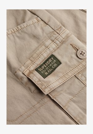 Beige stof med en glat tekstur, med en syet lomme og et grønt mærke, der siger "SUPERDRY UTILITY WEAR M.F.G. CO."