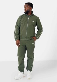 Olijfgroene zip-up jack en bijpassende broek met witte accenten. De stof lijkt lichtgewicht; de outfit wordt gecombineerd met witte sportieve schoenen.