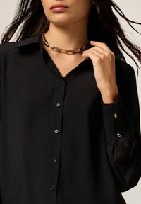 Black button-up shirt with a smooth texture, featuring a classic collar and decorative cuff buttons. Worn with a chain link necklace.