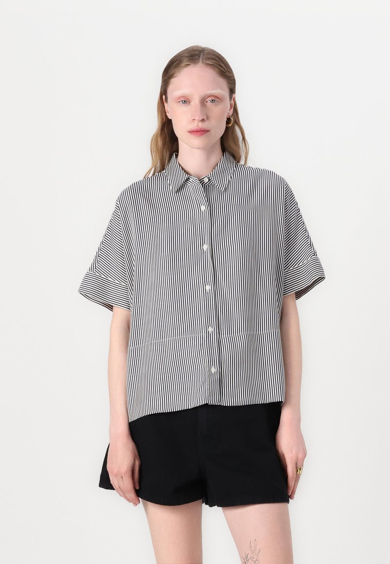 Camisa de manga corta a rayas en blanco y negro con botones en la parte delantera, que presenta un cuello clásico y un corte holgado, combinada con pantalones cortos negros.