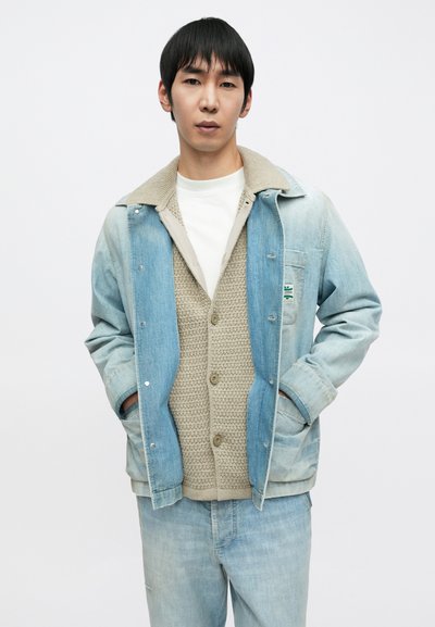 Τζιν μπουφάν σε ανοιχτό μπλε χρώμα με μπεζ, υφασμένο πουλόβερ από κάτω. Διαθέτει μεγάλα κουμπιά, γιακά και μπροστινές τσέπες.