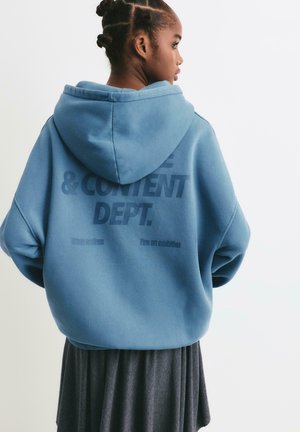 Jongere met ingevlochten haar die een blauwe oversized hoodie met tekst op de achterkant draagt, in combinatie met een donkere plissé rok.