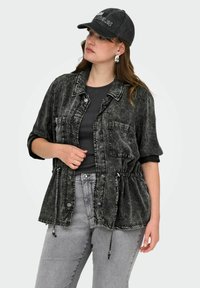 Črna denim jakna s sproščenim krojem, kratkimi rokavi, nastavljivim vrvico pri pasu in kontrastnim šivanjem. V kombinaciji s sivimi kavbojkami in kapo.