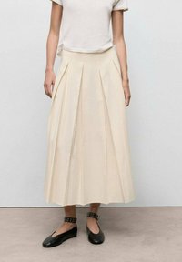 Pleated beige midi skirt made from light fabric, featuring a high waistband and side pockets. Paired with black flat shoes.